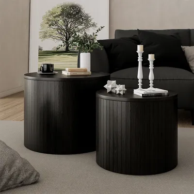 Two round black coffee tables with ribbed sides are placed on a gray rug in a living room setting. A painting of a tree hangs on the wall behind the tables, and a sofa with black cushions is visible in the background.