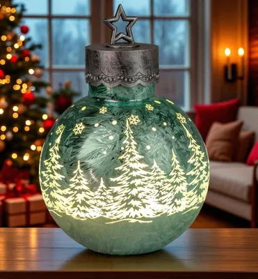 A decorative LED glass ball with a Christmas tree motif, placed on a wooden table in front of a Christmas tree and a living room setting.