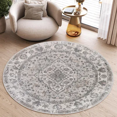 A round, vintage-style rug with a distressed pattern in shades of gray and white is placed on a wooden floor in a living room setting. A modern, light-colored armchair with cushions sits to the left of the rug, and a small, gold-toned side table with a boo