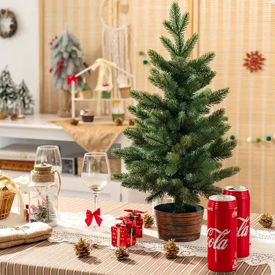 A small artificial Christmas tree is set up on a table with festive decorations, including gift boxes, pinecones, and Coca-Cola cans.