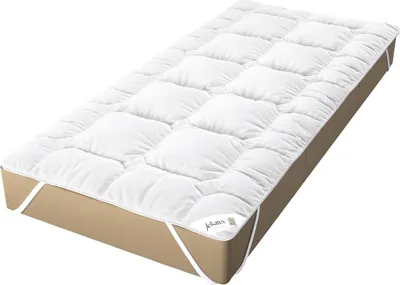 A white mattress topper with a quilted pattern is placed on top of a tan mattress. White elastic straps are visible at the corners of the topper, designed to secure it to the mattress.
