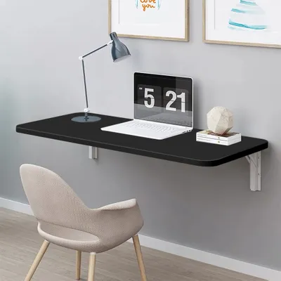 A black wall-mounted folding desk with a laptop, lamp, and decorative object on it, with a chair in front.