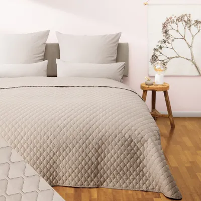 A neatly made bed with a beige quilted bedspread and pillows, in a room with a pink wall and a wooden stool with a lamp.
