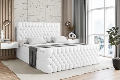 A white tufted boxspring bed with a headboard and mattress is shown in a bedroom setting.