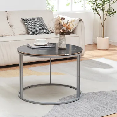 A round coffee table with a marble top and metal legs is placed on a rug in front of a sofa. A vase with dried flowers and a book with a cup of tea are on the table.