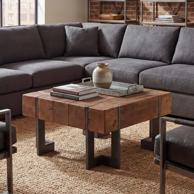 A square wooden coffee table with metal legs is placed in front of a large sectional sofa in a living room setting. Books and a decorative tray with a vase are on the table.