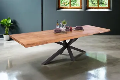 A wooden dining table with a live edge and a black metal spider leg base is set in a modern dining room with a dark gray wall and a concrete floor. Two windows with greenery visible are in the background.