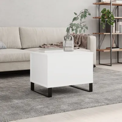 A white rectangular coffee table with black metal legs is placed on a gray rug in a living room setting. A sofa is visible in the background, along with a bookshelf.