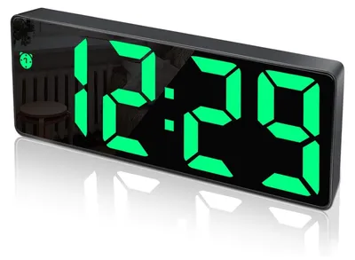 A digital alarm clock with a black casing and green LED display showing the time 12:29. The clock has a reflective surface and is shown on a reflective surface, casting a shadow.