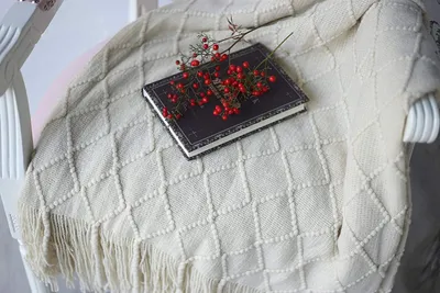 A beige knitted blanket draped over a white chair, with a book and red berries on top.