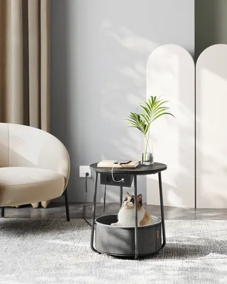 A cat is sitting in a round bed integrated into a side table, with a book and phone on top of the table.