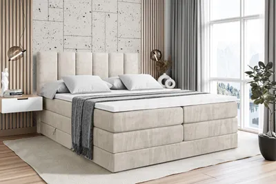 A bedroom scene featuring a large box spring bed with a headboard, mattress, and topper. The room has modern decor with textured walls, wooden accents, and a large window with curtains.