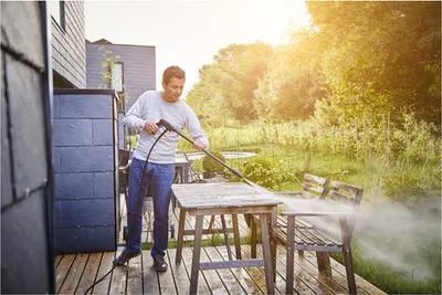 A man is pressure washing an outdoor wooden table and chair set on a wooden deck.