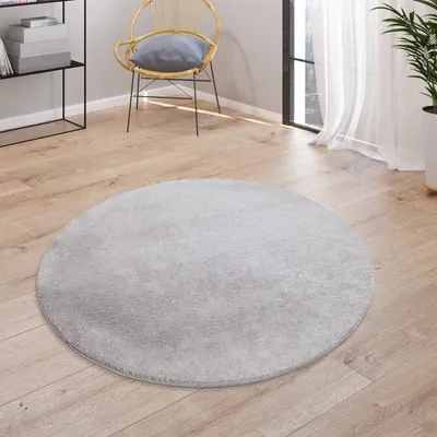 A round, gray rug is placed on a wooden floor in a room with a chair and a shelf.