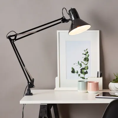 A black desk lamp is clamped to a white desk, with a framed picture and two small cups behind it.