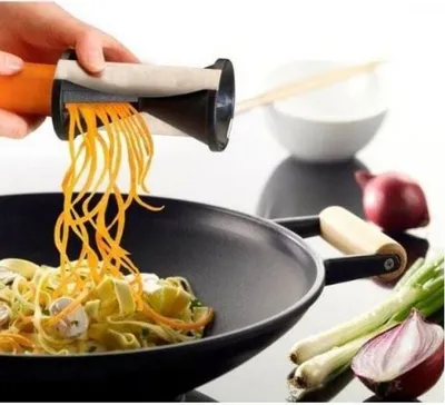 A hand is using a spiralizer to cut carrots into noodles over a wok filled with pasta and vegetables. Other vegetables like onions and scallions are visible in the background.