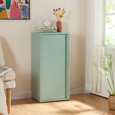 A mint green metal cabinet with a sliding door stands in a room next to a chair and a window with sheer curtains. Decorative items are placed on top of the cabinet.