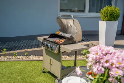A gas grill is set up on a patio with food cooking on it, and flowers in the foreground.