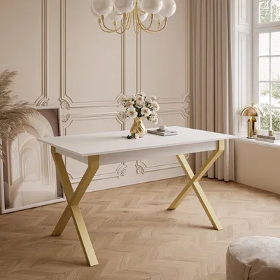 A white dining table with gold X-shaped legs is set in a well-lit room with decorative molding on the walls and a large window with beige curtains.