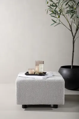 A light gray, textured ottoman sits in front of a light gray wall. A plant in a black pot is to the right of the ottoman. Two candles and a book are on top of the ottoman.