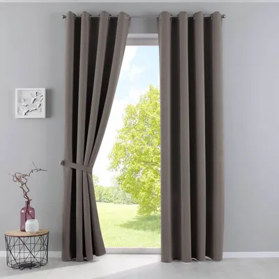 A pair of taupe curtains are shown tied back, revealing a window with a green tree and blue sky outside. A small side table with a vase and a decorative object is to the left.