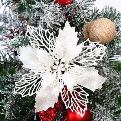 A close-up view of a white glitter poinsettia ornament on a Christmas tree, surrounded by pine needles, red berries, and a small burlap sack ornament.