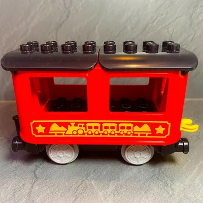 A red LEGO DUPLO train car with black roof and wheels sits on a gray surface.