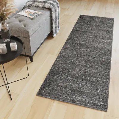 A dark gray runner rug is placed on a light wood floor next to a gray tufted storage bench with a blanket draped over it and a black side table with candles and a vase.
