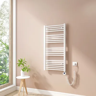 A white electric towel radiator mounted on a light pink wall next to a window.