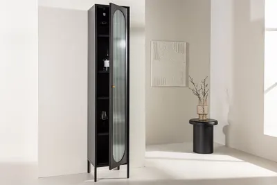 A tall, narrow black cabinet with a glass door is shown in a room with a small round table and artwork on the wall.