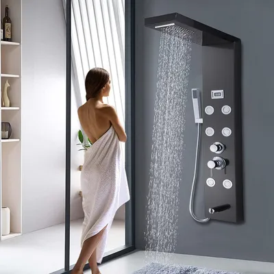 A woman wrapped in a towel stands in front of a modern shower panel as water cascades down.
