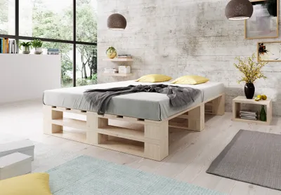 A bedroom scene featuring a pallet bed with bedding and pillows, a bedside table with a vase of flowers, and a large window overlooking greenery.