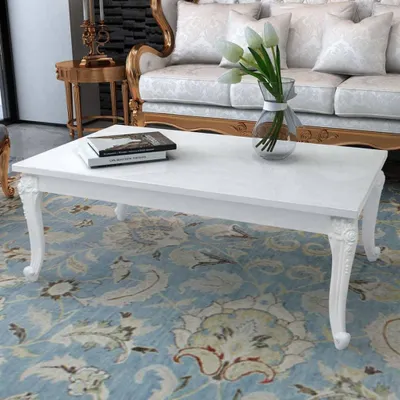 A white coffee table with books and a vase of flowers on it, placed on a patterned rug in front of a sofa.