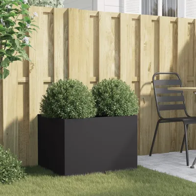 A black rectangular planter with two round green bushes sits on a grassy area in front of a wooden fence. A metal chair and table are visible to the right.