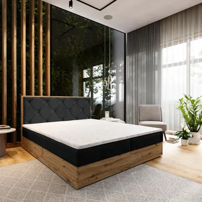 A bedroom scene featuring a large bed with a tufted headboard, a wooden base, and a white mattress and topper. The room has large windows with sheer curtains, wooden wall paneling, and a potted plant.