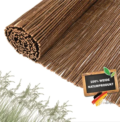 A rolled-up privacy screen made of willow branches is shown diagonally across a white background, with some grass in the lower left corner. A small chalkboard with text and the German flag is in the lower right.