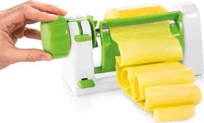 A hand is shown turning the crank of a vegetable slicer, producing long, thin strips of yellow vegetable.