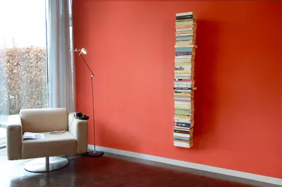 A modern living room with a chair, floor lamp, and a tall, narrow bookshelf filled with books mounted on a coral wall.