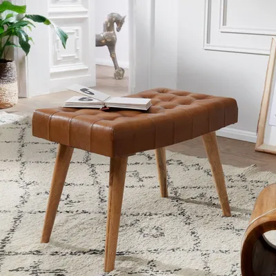 A brown leather tufted ottoman with wooden legs sits on a patterned rug in a room.