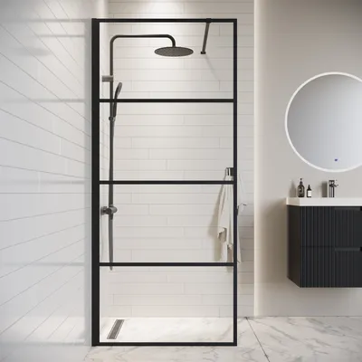 A modern bathroom with a black-framed glass shower enclosure, a shower head, and a towel hanging on the side. A vanity with a round mirror is visible in the background.