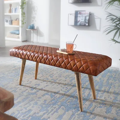 A brown quilted leather bench with wooden legs sits on a patterned rug in a well-lit room.