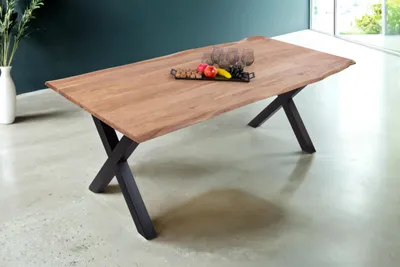 A wooden dining table with a live edge and black X-shaped metal legs, set in a modern dining room with a vase of greenery and fruit on the table.