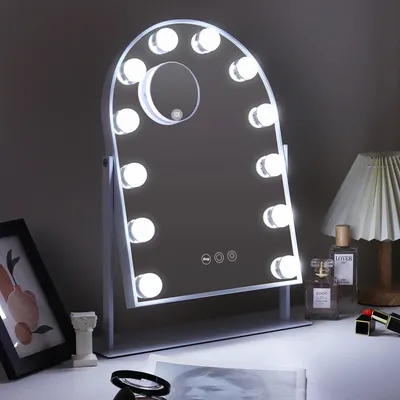 A vanity mirror with lights is displayed on a desk, with some decorative items and perfume bottles in the background.