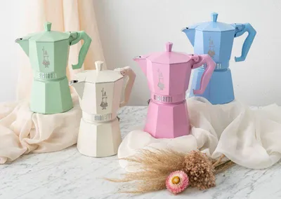 Four Bialetti Moka Express coffee makers in pastel colors (green, cream, pink, blue) arranged on a surface with draped fabric and dried flowers.
