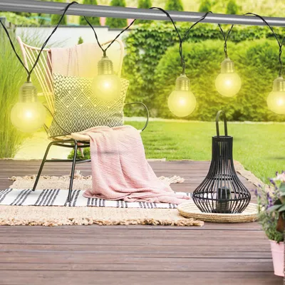 A string of solar-powered LED lights hangs over a patio chair with a blanket and pillow, set on a wooden deck with a lantern and plants.