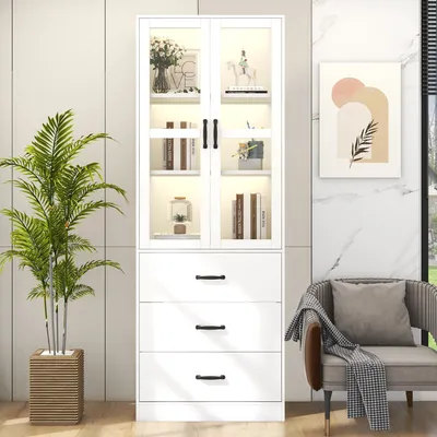 A white display cabinet with glass doors and three drawers, placed next to a chair with a blanket and a potted plant.