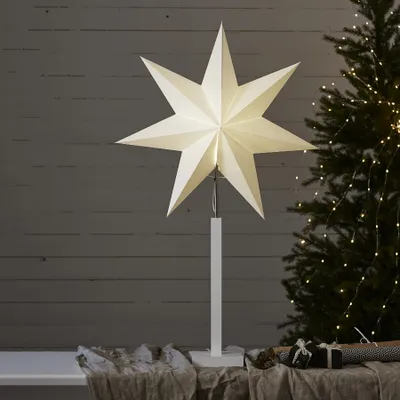 A white star-shaped lamp on a stand, placed in front of a wooden wall and a Christmas tree with lights.