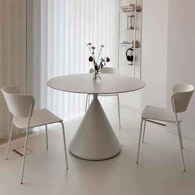 A minimalist dining set with a round table and two chairs in a bright, white room with vertical blinds.