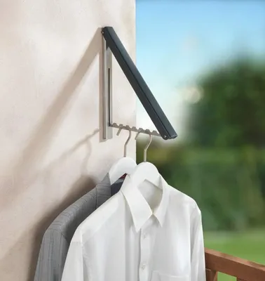 A wall-mounted folding clothes rack with two shirts hanging on it, outdoors with greenery and sky in the background.