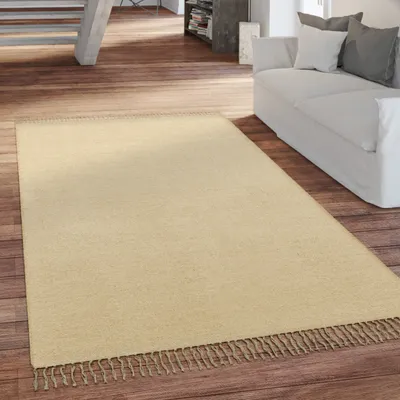 A beige rug with fringes is placed on a wooden floor in a living room setting, with a white sofa visible in the background.